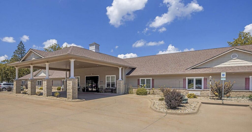 A single-story building with a covered entrance supported by stone columns, surrounded by a paved driveway, small shrubs, and a partly cloudy sky overhead.