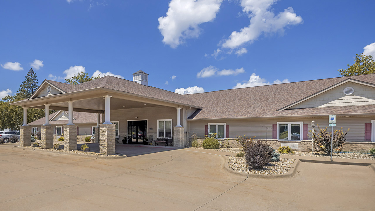 Single-story building with a large covered entrance supported by columns, surrounded by a paved driveway, small landscaped areas, and shrubs under a blue sky with scattered clouds.
