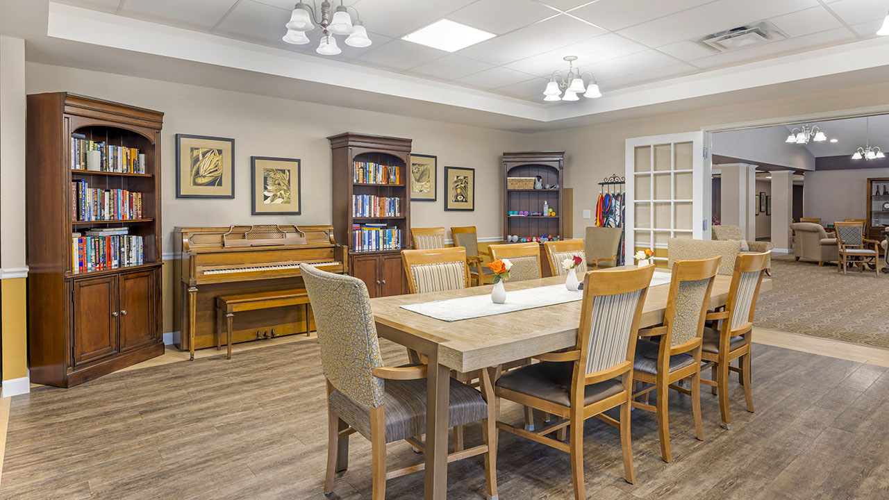 A spacious, well-lit room with a large wooden dining table surrounded by chairs, bookshelves filled with books, framed art on the walls, a piano, and soft overhead lighting. Another room is visible through open doors.