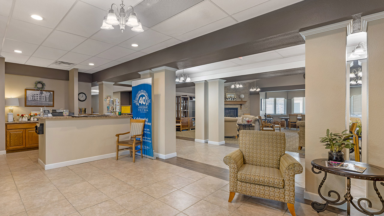 A spacious, well-lit lobby with tiled floors, armchairs, a reception desk, and a blue welcome sign. The area features neutral tones and leads into a cozy lounge with tables, chairs, and a fireplace in the background.