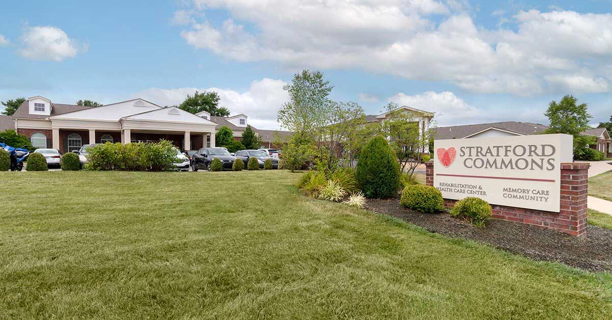 A wide view of Stratford Commons, a premier Memory Care and rehabilitation center. The single-story building boasts a driveway lined with parked cars, while the grassy front yard showcases neatly trimmed bushes and a sign proudly displaying the facility's name.