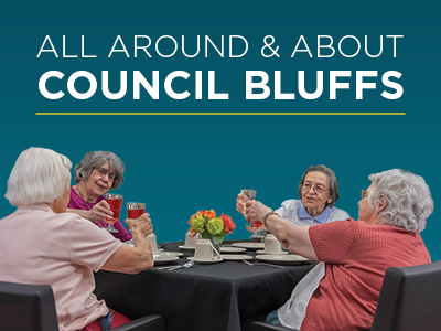 Four elderly women sit around a table, raising glasses in a toast, with flowers and dishes in front of them. The text above reads, “All Around & About Council Bluffs.”.