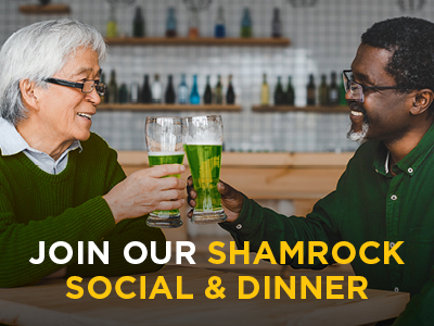 Two men smiling and clinking glasses of green beer in a bar. Both are wearing green sweaters. Text overlay reads, 