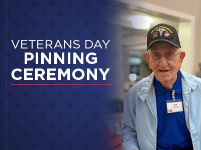 Elderly man in a military cap and light blue jacket smiles, standing beside a sign that reads 
