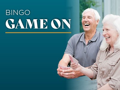 Two smiling older adults holding bingo cards sit at a table, enjoying themselves. The words 