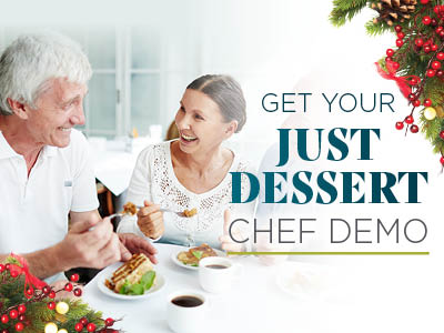 An older man and woman smile and enjoy dessert together at a table in a Senior Living Community, with festive holiday decorations in the corners. Text reads, 