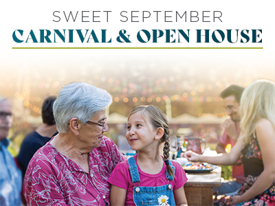 An older woman and a young girl smile at each other at an outdoor event. People sit and talk at tables in the background. Text above reads: “Sweet September Carnival & Open House.”.