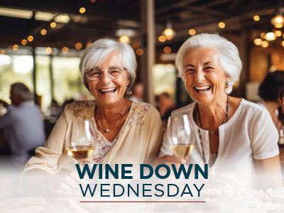 Two elderly women with white hair smile warmly while holding glasses of white wine in a cozy, warmly lit restaurant. The text overlay at the bottom reads 