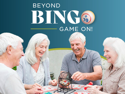Four smiling older adults sit around a table playing bingo. They are holding cards and using a bingo cage with balls. The text reads, “Beyond Bingo. Game On!” on a blue background.