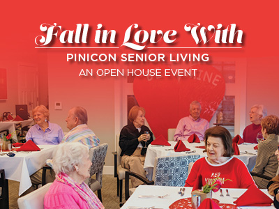 A group of seniors sit at decorated tables, smiling and socializing at a dining area for a Valentine’s-themed “Fall in Love With Pinicon Senior Living” open house event.