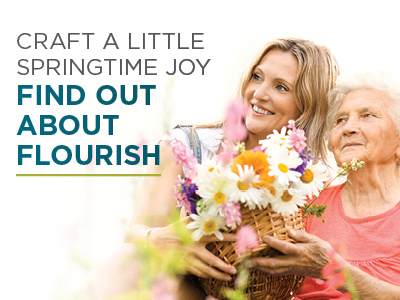 A young woman and an elderly woman smile together outdoors, holding a basket of colorful spring flowers. Text reads: