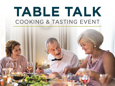 A group of people sits around a table set with food and drinks, smiling and conversing. The text above reads 