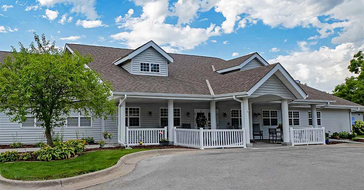 A single-story gray house with a gabled roof, white trim, and a covered porch with white railings. A small tree and landscaped shrubs are in the front yard, and the sky is partly cloudy.