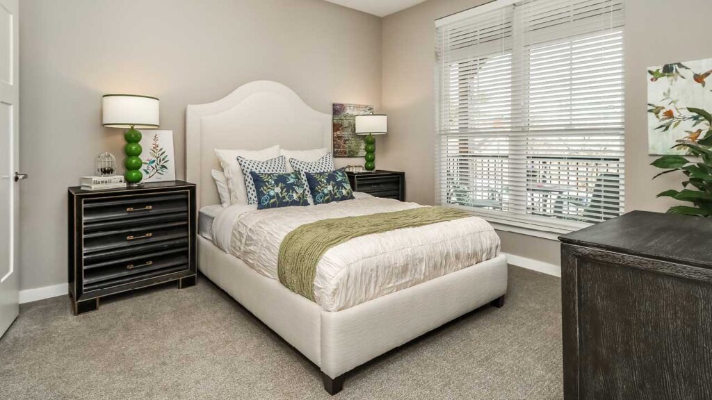 A well-lit senior living bedroom with a large bed in the center, adorned with white and light blue bedding and green accent cushions. There are two black nightstands with green lamps on each side of the bed. A large window with blinds is on the right side of the room.