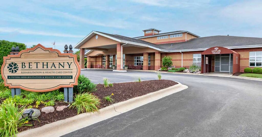 Exterior view of Bethany Rehabilitation & Health Care Center, a brick building with a covered entrance, sign, well-maintained landscaping, and driveway under a blue sky.
