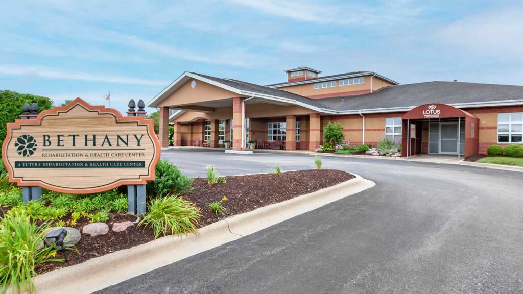 A large Rehabilitation & Health Care Center with a covered entrance and a sign reading "Bethany Rehabilitation & Health Care Center" stands beside a landscaped driveway under a partly cloudy sky.