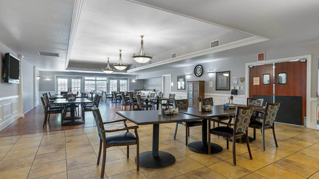 A spacious, well-lit dining room with multiple tables and chairs, large windows at the back, tiled floors in the foreground, wood floors in the background, and modern light fixtures on the ceiling.