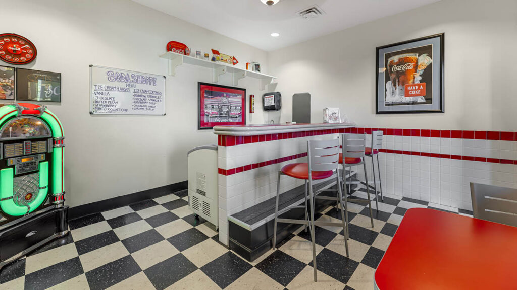 A small retro diner with black-and-white checkered floor, red and white tiled counter, gray chairs, a neon jukebox, and Coca-Cola decor on the walls. Shelves hold various vintage items.