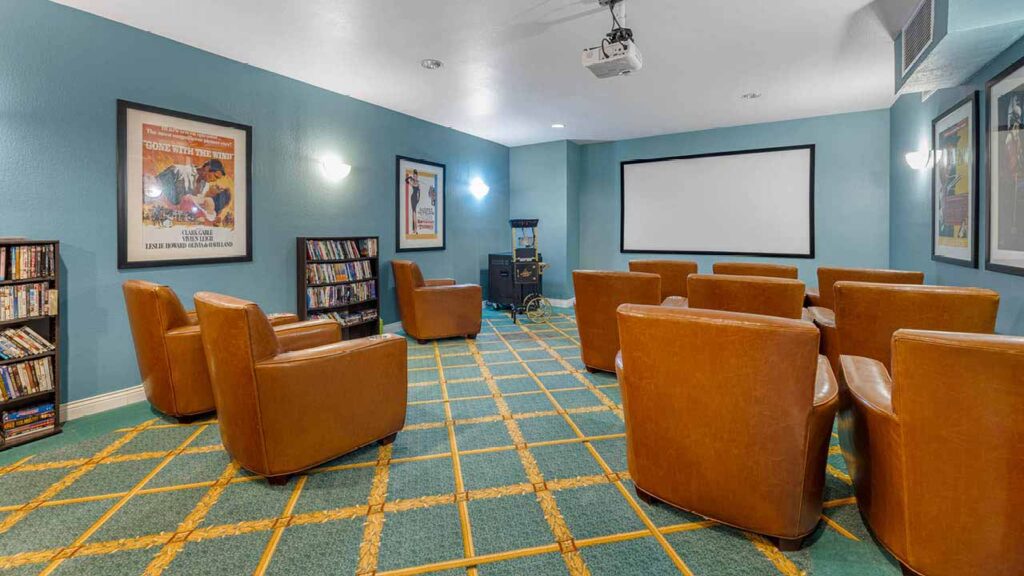 A small home theater room with brown leather chairs facing a large screen, blue walls, movie posters, DVD shelves, a popcorn machine, and wall-mounted lights.