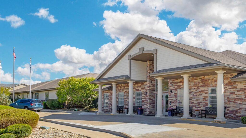 A single-story building with brick and white siding, columns at the entrance, benches near the doorway, and a car parked outside, under a bright blue sky with scattered clouds.