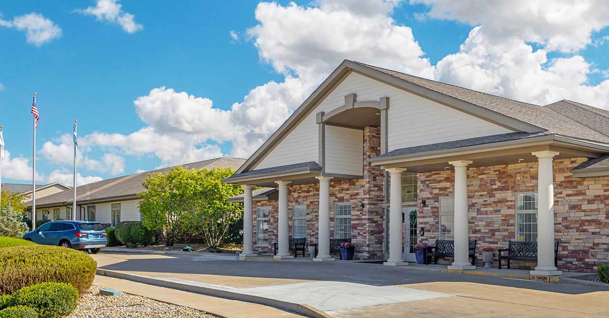 A single-story building with a brick and stone exterior, white columns, and a covered entrance. Several benches and a few cars are visible, with trees and an American flag nearby under a partly cloudy sky.