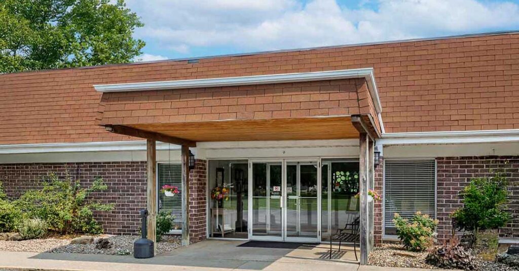 A single-story brick Rehabilitation & Health Care Center with a brown shingled roof and a covered entrance. Glass double doors are at the center, flanked by flower pots and greenery. A chair sits near the entryway.