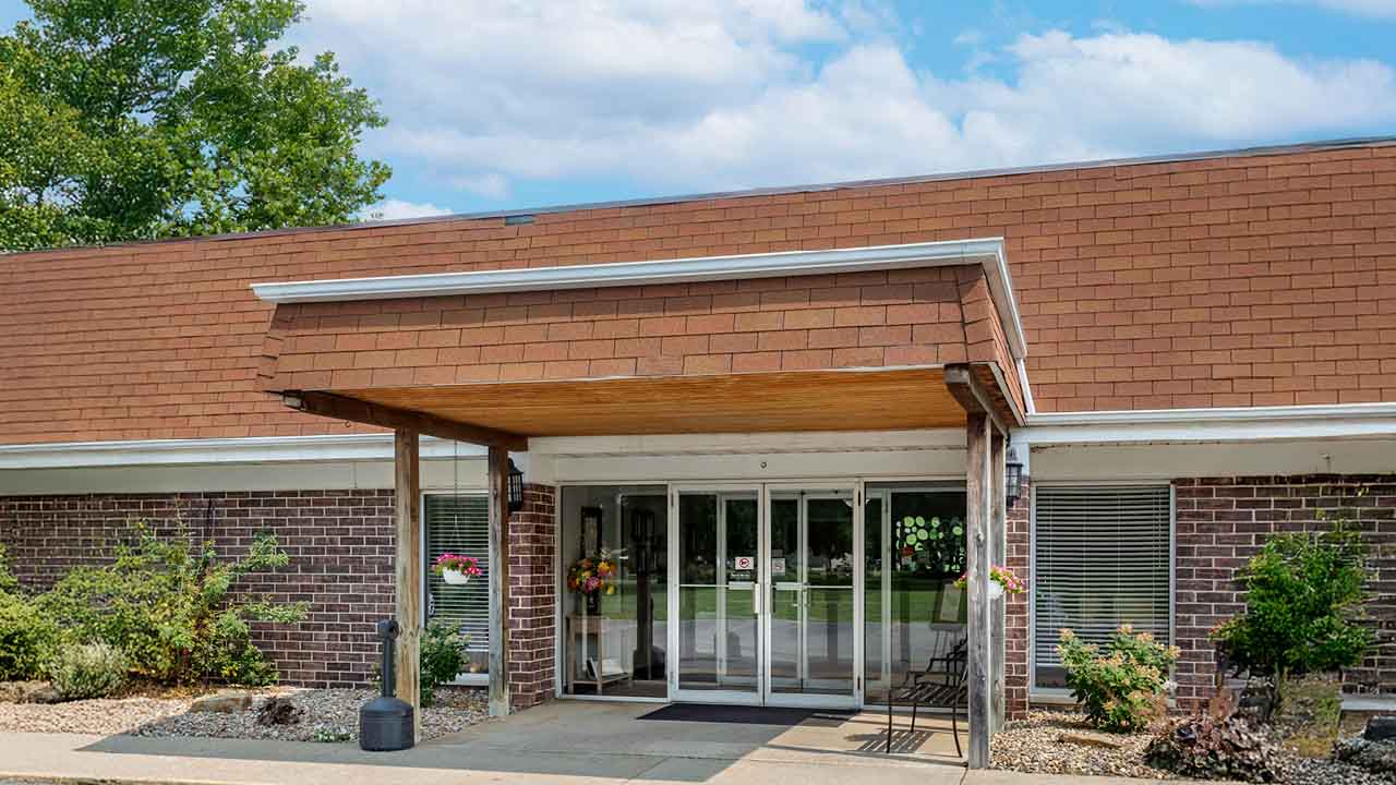 A single-story brick Rehabilitation & Health Care Center with a brown shingled roof and covered entrance. Glass doors and windows reflect greenery, while potted plants, flowers, and chairs create a welcoming entryway.
