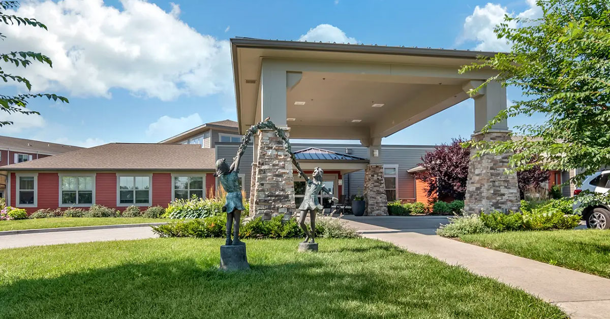 The exterior view of the Rehabilitation & Health Care Center features a large covered entrance. Two statues of children playing adorn the grassy area in front, surrounded by trees and shrubs. The sky is a crisp blue with scattered clouds, casting a serene atmosphere over this inviting space.