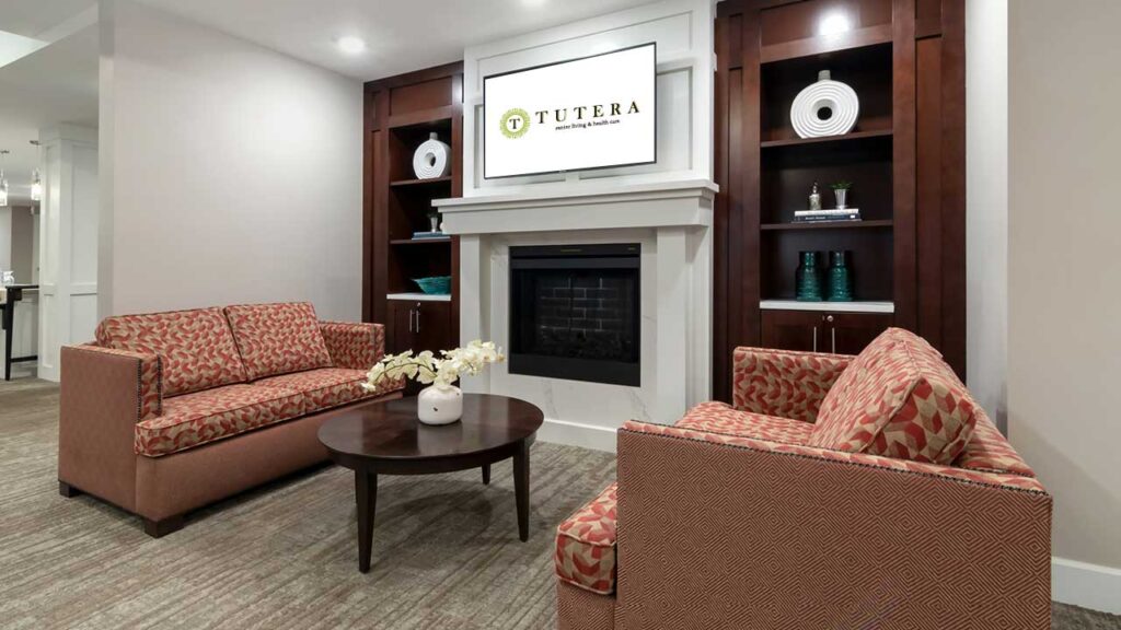 A cozy senior living lounge area featuring two patterned red sofas facing each other with a dark wooden round table in between. A fireplace is centered on the back wall, flanked by two wooden bookshelves. A flat-screen TV displaying 