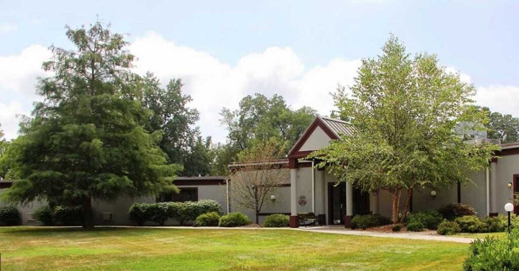 A single-story modern building with a peaked roof entryway, surrounded by green trees, shrubs, and a well-kept grassy lawn under a partly cloudy sky.