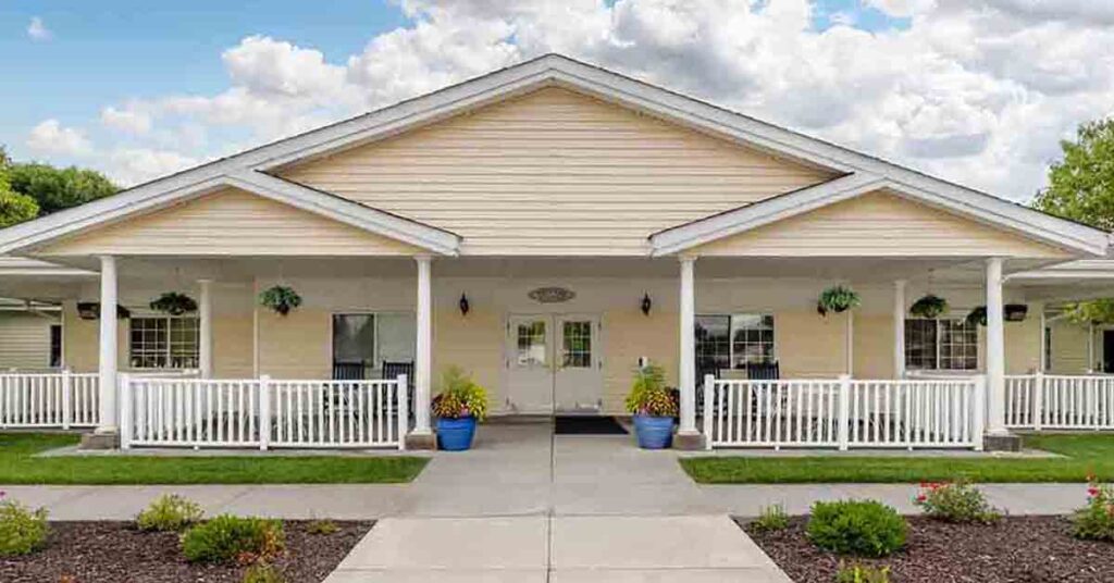 Single-story building with cream siding, a covered porch, white railings, and potted plants by the entrance; neatly trimmed shrubs and a walkway lead to double doors under a partly cloudy sky.