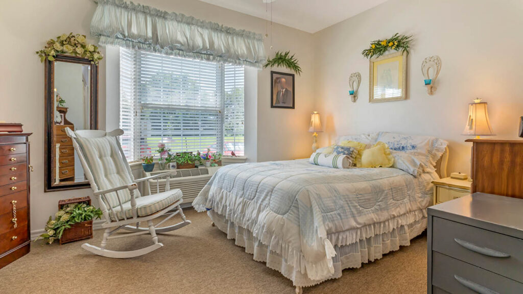 A cozy bedroom in Assisted Living Muskogee features a large bed with white ruffled bedding, a wooden rocking chair, potted plants by a sunny window, dressers, lamps, and decorative wall accents for a warm, inviting atmosphere.
