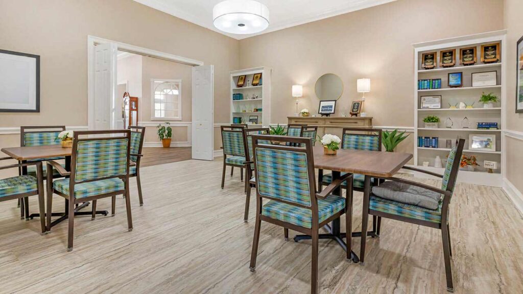 A brightly lit Assisted Living Muskogee room with tables and blue-green patterned chairs, light wood flooring, built-in shelves with books and awards, a sideboard with lamps and decor, and double doors leading to another area.