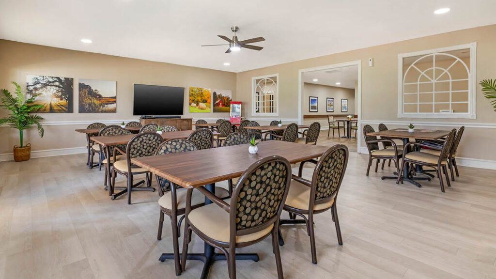 A bright, spacious Assisted Living Muskogee room with wooden tables and patterned chairs arranged in rows, wall art, a large TV, ceiling fan, potted plants, and a view into an adjoining room through large open windows.