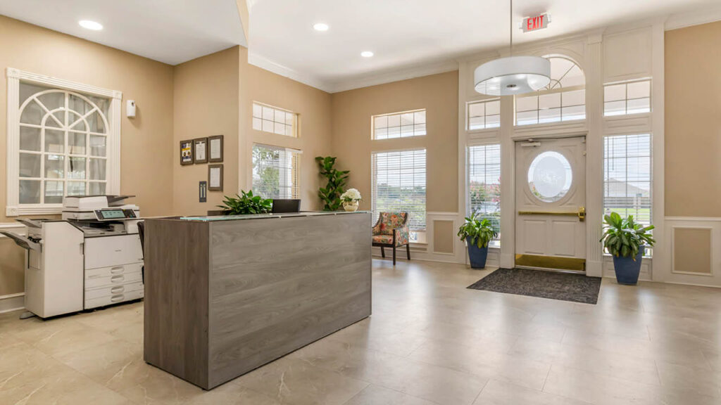 A bright, welcoming office lobby for Assisted Living Muskogee features beige walls, large windows, a wooden reception desk with plants, a printer, two chairs by the door, and sunlight streaming in from outside.