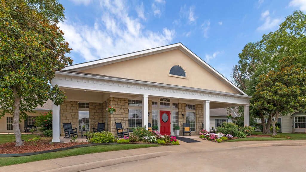 Single-story assisted living building with vibrant floral landscaping and a decorative red door at Country Gardens in Muskogee, OK, Muskogee County.