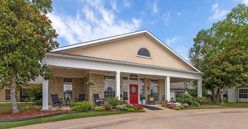 Large, single-story Assisted Living Muskogee building with a columned entrance, tan stone facade, red front door, rocking chairs on the porch, and landscaped flower beds. Trees and blue sky are visible in the background.