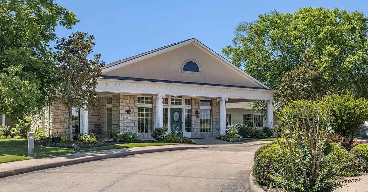 Single-story Assisted Living Muskogee building with a covered entrance, stone and siding exterior, white columns, and large windows, surrounded by green trees and shrubs on a sunny day. A curved driveway leads to the entrance.