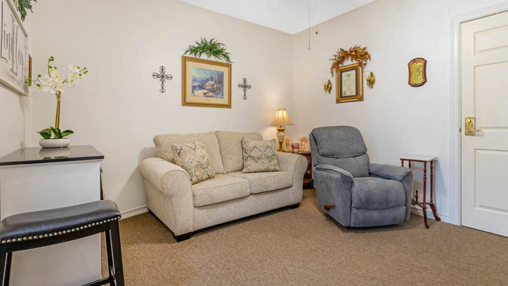 A cozy living room in Assisted Living Muskogee features a beige loveseat, gray recliner, side table with lamp, and framed art on the walls. Neutral carpeting and a small counter with a stool complete this inviting space.