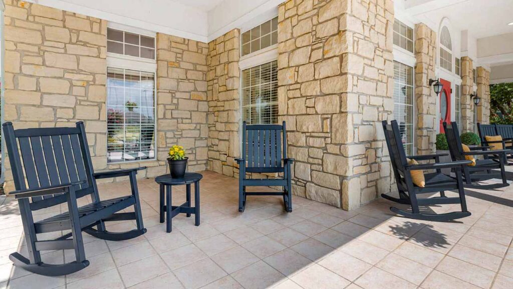 A stone porch at Assisted Living Muskogee features three dark wooden rocking chairs and a small round table with a yellow potted plant, set against windows with white blinds.