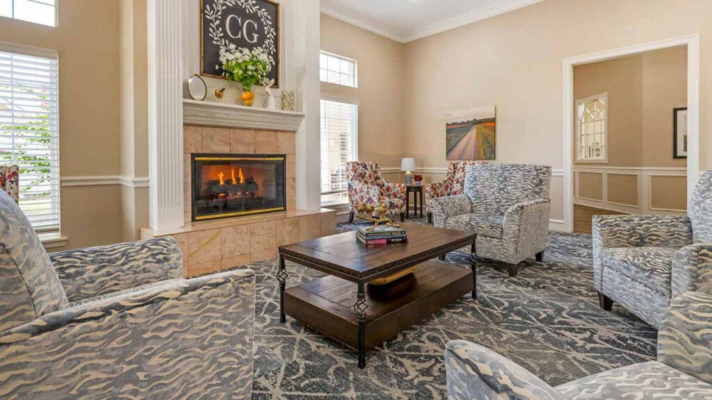A cozy living room in Assisted Living Muskogee features a fireplace, four patterned armchairs, a wooden coffee table, large windows with blinds, and wall art above the mantel. Light neutral walls and a patterned rug complete the space.