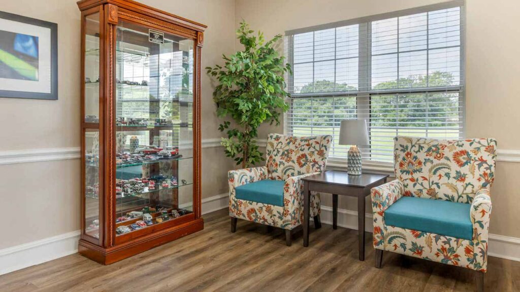 A cozy corner in Assisted Living Muskogee with two floral armchairs and a small table between them, set near large windows. A tall glass display cabinet filled with collectibles and a leafy plant stand nearby on wood flooring.