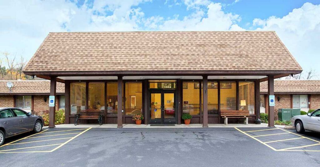Single-story brick Rehabilitation & Health Care Center with a large shingled roof, glass entrance doors, and covered walkway. Two benches and potted plants are in front, with cars parked in accessible spaces by the entrance.