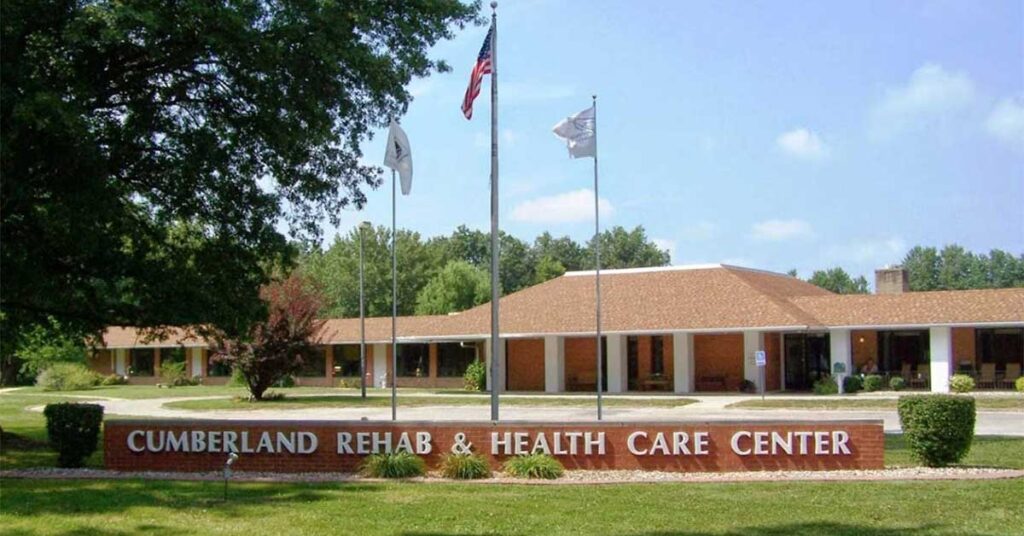 A brick sign reading “Cumberland Rehab & Health Care Center” stands in front of a large, single-story building with a flagpole and three flags, surrounded by grass, trees, and flowering shrubs.