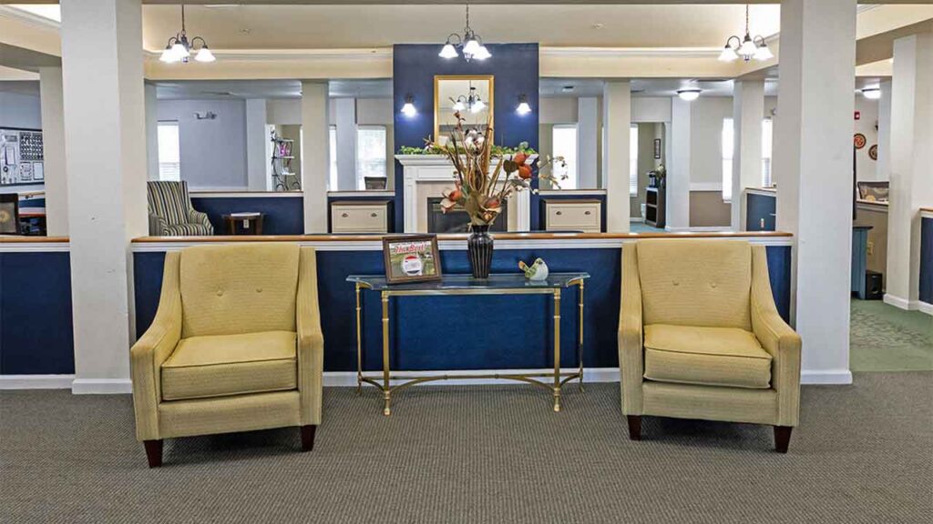 A cozy seating area featuring two mustard-yellow armchairs flanking a glass-topped console table with a vase of flowers. The background has dark blue panels, shelves with assorted items, wall lights, and overhead lighting fixtures.