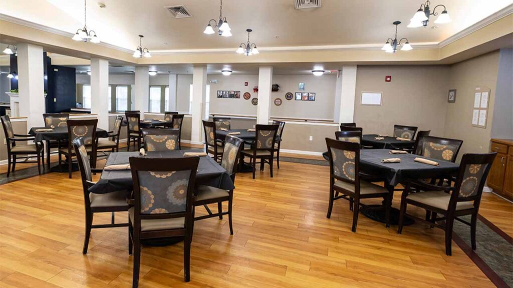 A well-lit dining room with wooden flooring, neatly arranged tables covered with black tablecloths, and chairs with patterned upholstery. The walls are adorned with framed art and decorative plates. Overhead lights and windows illuminate the space.
