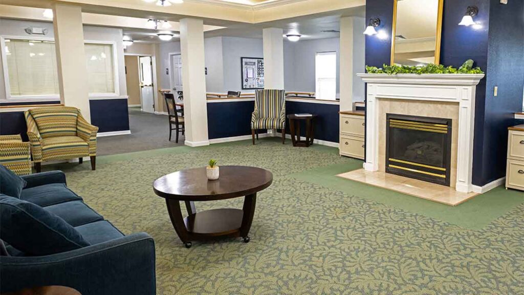 A spacious and well-lit living room with green patterned carpet, dark blue and white walls, and a large fireplace with a white mantel. The room features comfortable seating, including a dark blue sofa, a striped chair, two armchairs, and a wooden coffee table.