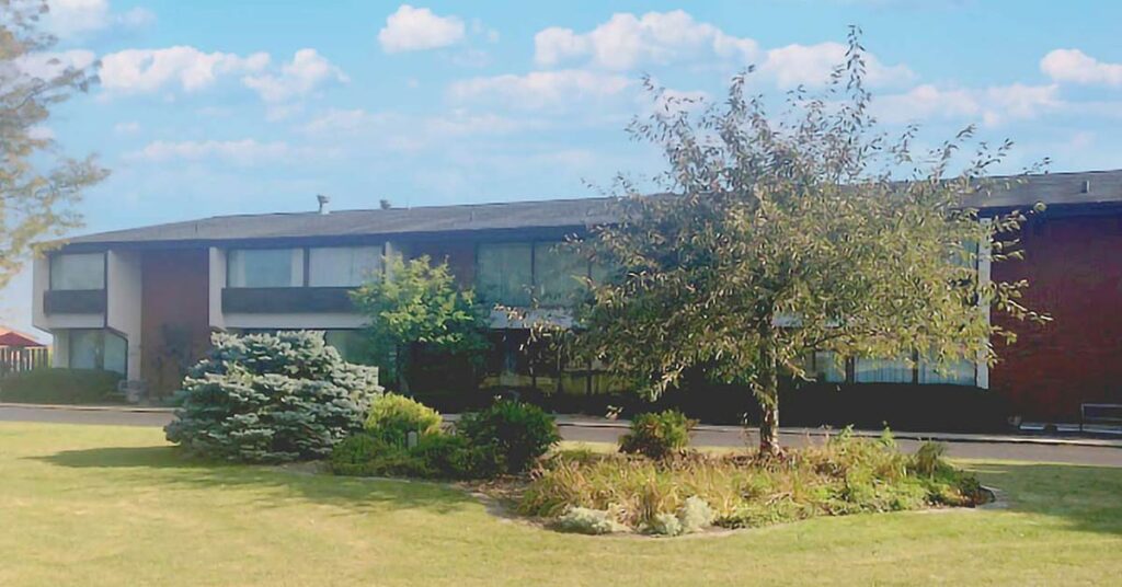 A two-story brick and glass building with large windows, surrounded by green grass, various shrubs, and a small tree, under a blue sky with scattered clouds.