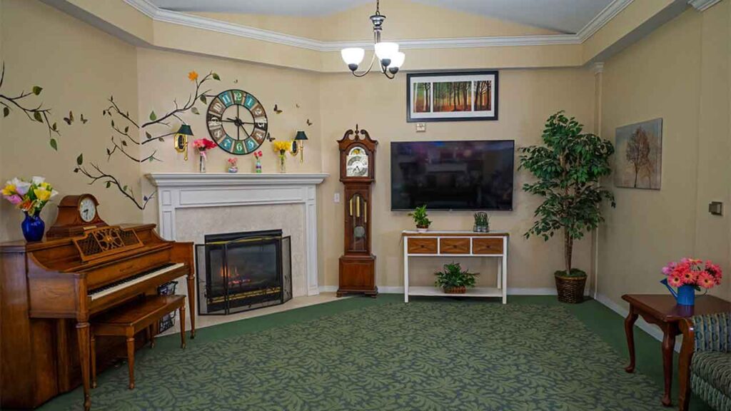 A cozy living room with a green patterned carpet, beige walls, and a white-trimmed ceiling. It features a piano, a fireplace, a grandfather clock, a flat-screen TV on a stand, and decorative plants. Wall art and flowers add a touch of color.