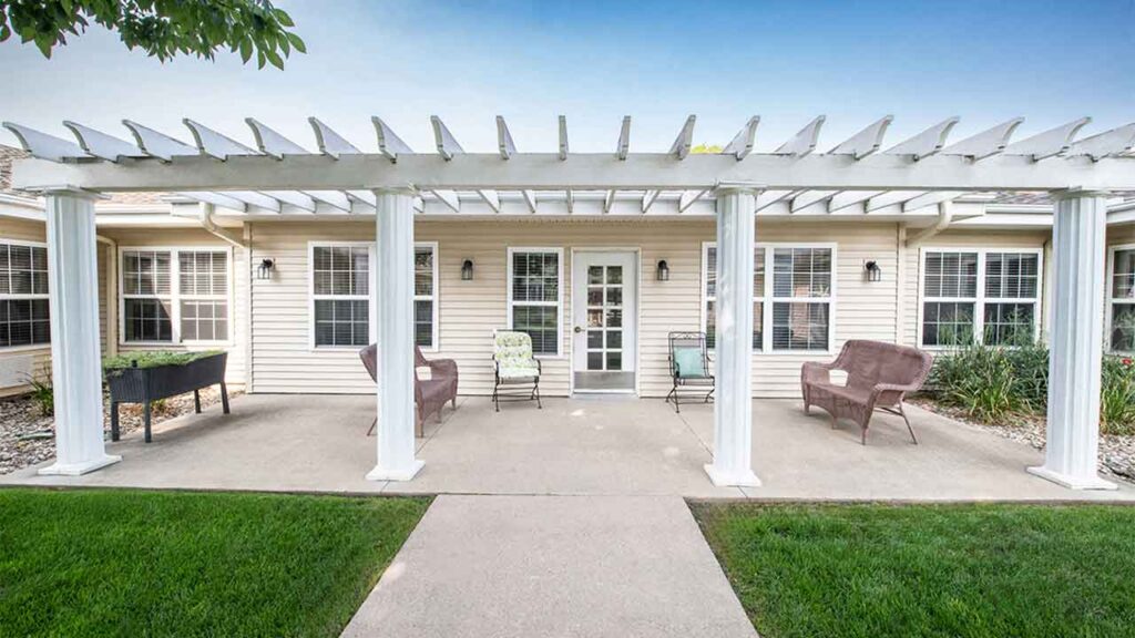 A patio with a white pergola extends from a beige house. The pergola is supported by four columns, and the patio is furnished with various outdoor seating, including chairs and a loveseat. Green grass and a tree are in the foreground, with a clear blue sky above.